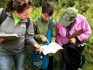 Three people stand together, outdoors, looking at an open book that the person in the center is holding.