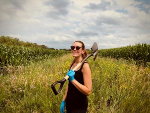 Corinn Rutkoski standing in a field holding a spade with a cloudy sky in the background