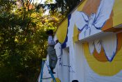 Image of a colorful mural being painted by artist standing on a ladder with trees in the background