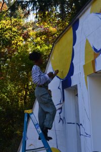 Image of artist Stephanie Clark painting a colorful mural while on a ladder with trees in the background.