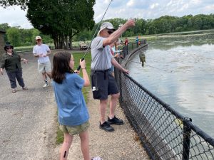 Image of people standing near fence at a lake holding fishing poles 