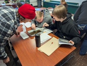 Image of 2 students at a table with papers, a tray of pond water, and sampling tools with teacher standing nearby