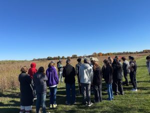 Image of a student group standing in the grass, facing towards a field, with a bright blue sky while listening to a talk. 